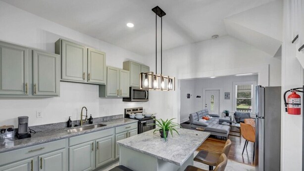 A modern kitchen with light green cabinets, a central island with bar stools, a microwave, and a stainless steel refrigerator. - The Scoreboard | Hot Tub (Columbia)
