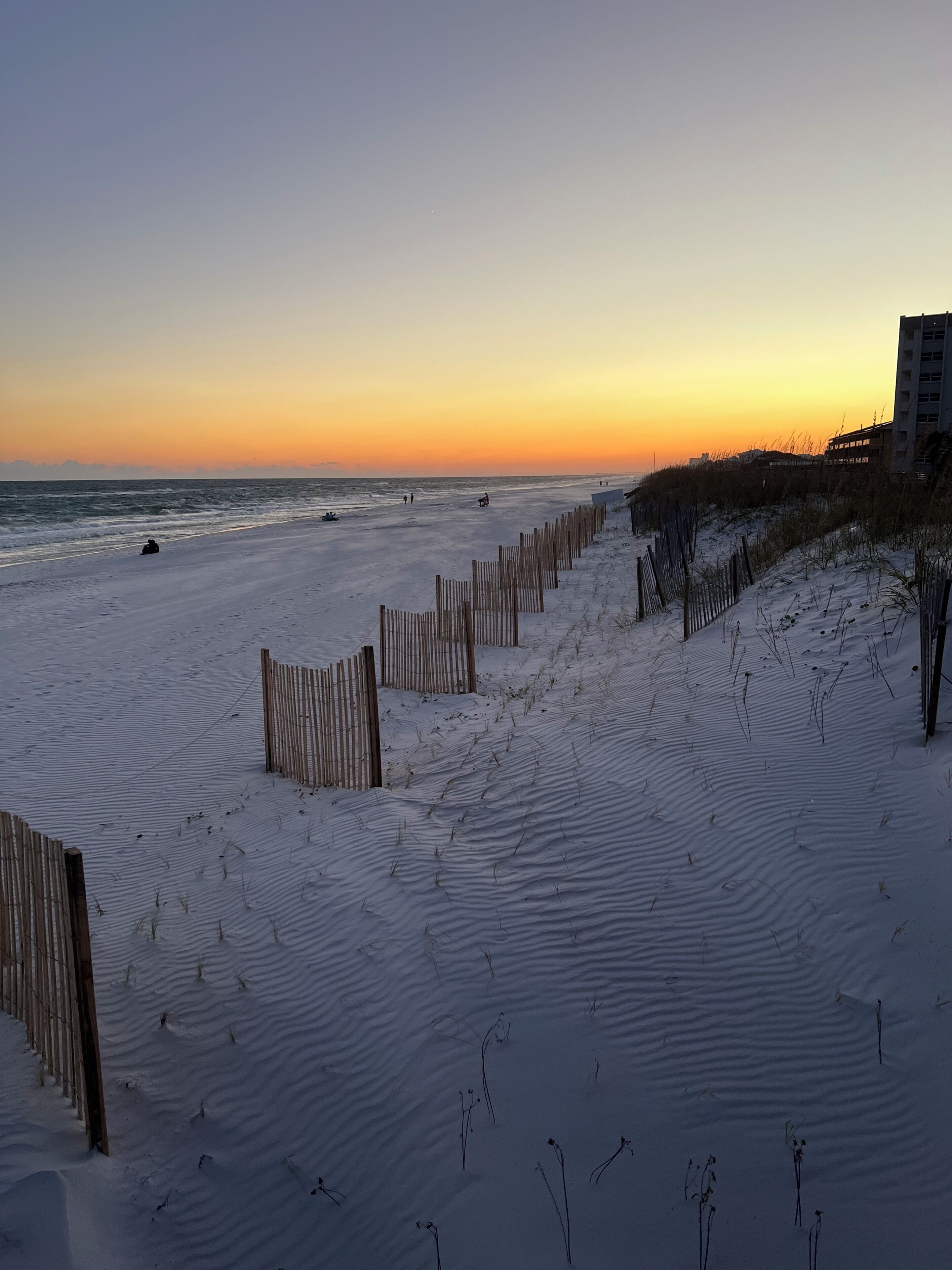 Ein Strand mit einer Holzbrüstung, Dünen und einem Gebäude im Hintergrund.