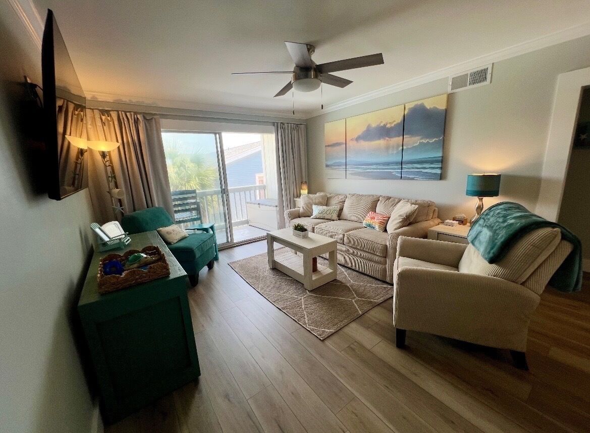 A living room with a sofa, armchairs, a coffee table, and a TV. There is a balcony with a view of the sea.