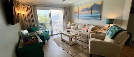 A living room with a sofa, armchairs, a coffee table, and a TV. There is a balcony with a view of the sea.