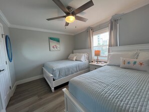 A bedroom with two beds, a ceiling fan, a nightstand with a lamp, and a beach-themed wall art.