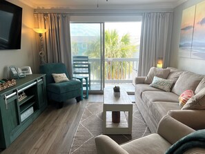 A living room with a sofa, armchair, coffee table, TV, and a balcony with a view.