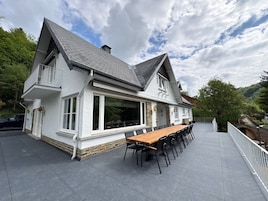 Una casa de dos pisos con un amplio patio, una mesa de comedor y asientos exteriores.