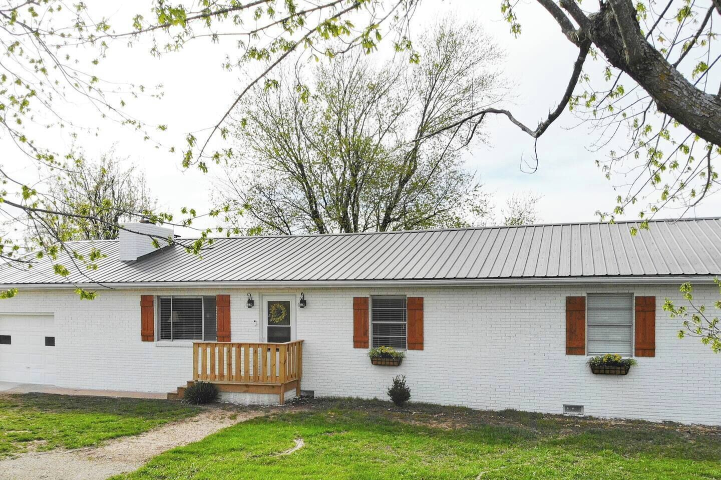 A single-story white brick house with a metal roof, wooden shutters, and a small porch with a bench.