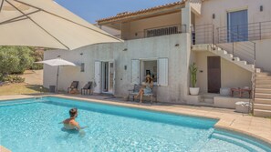 Ein Schwimmbecken mit einer Person im Wasser, eine Frau liest auf einer Bank, und eine Treppe führt zu einem Balkon.