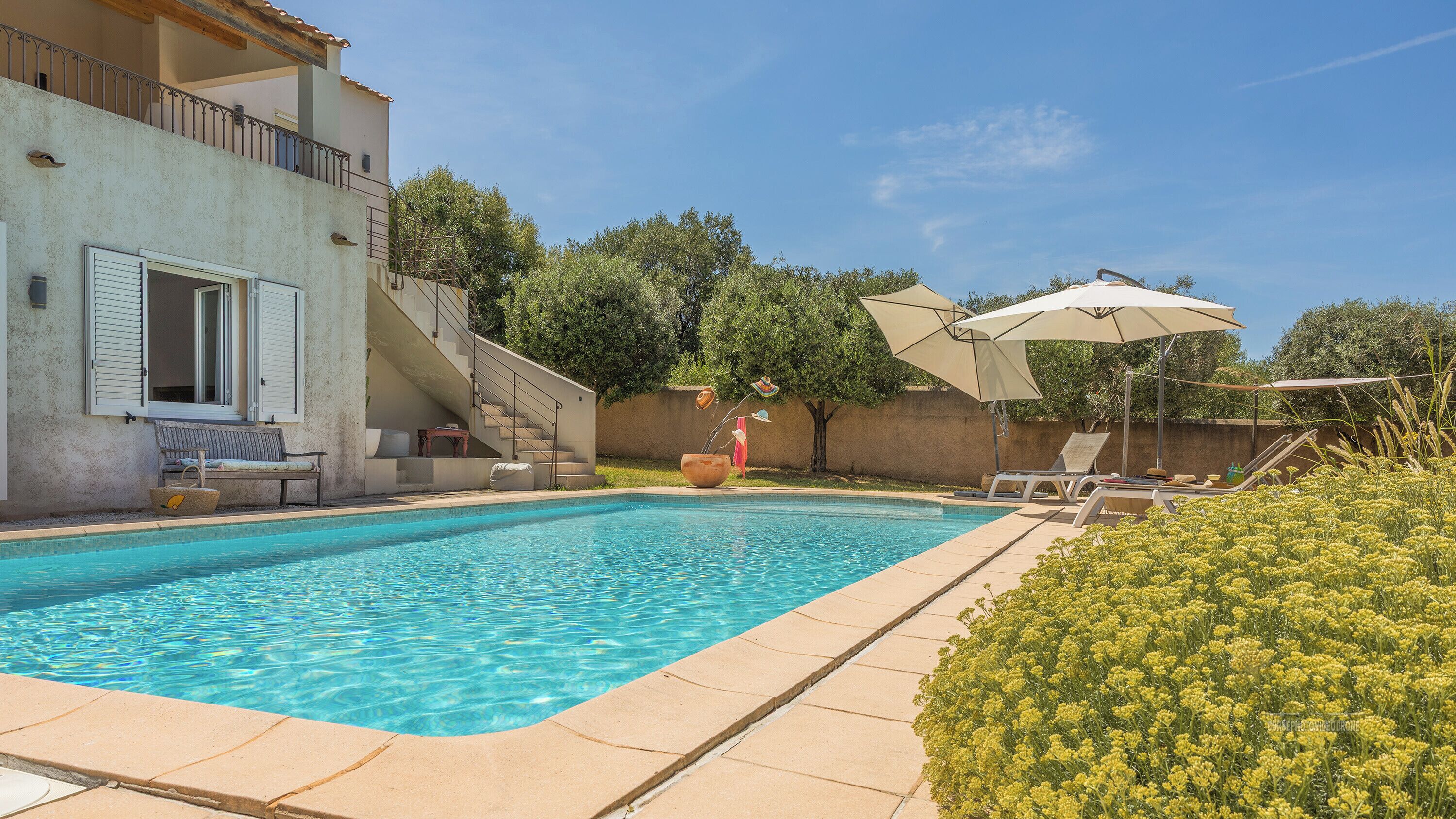 A swimming pool with a sun lounger and umbrella, a bench, and a staircase leading to a balcony.