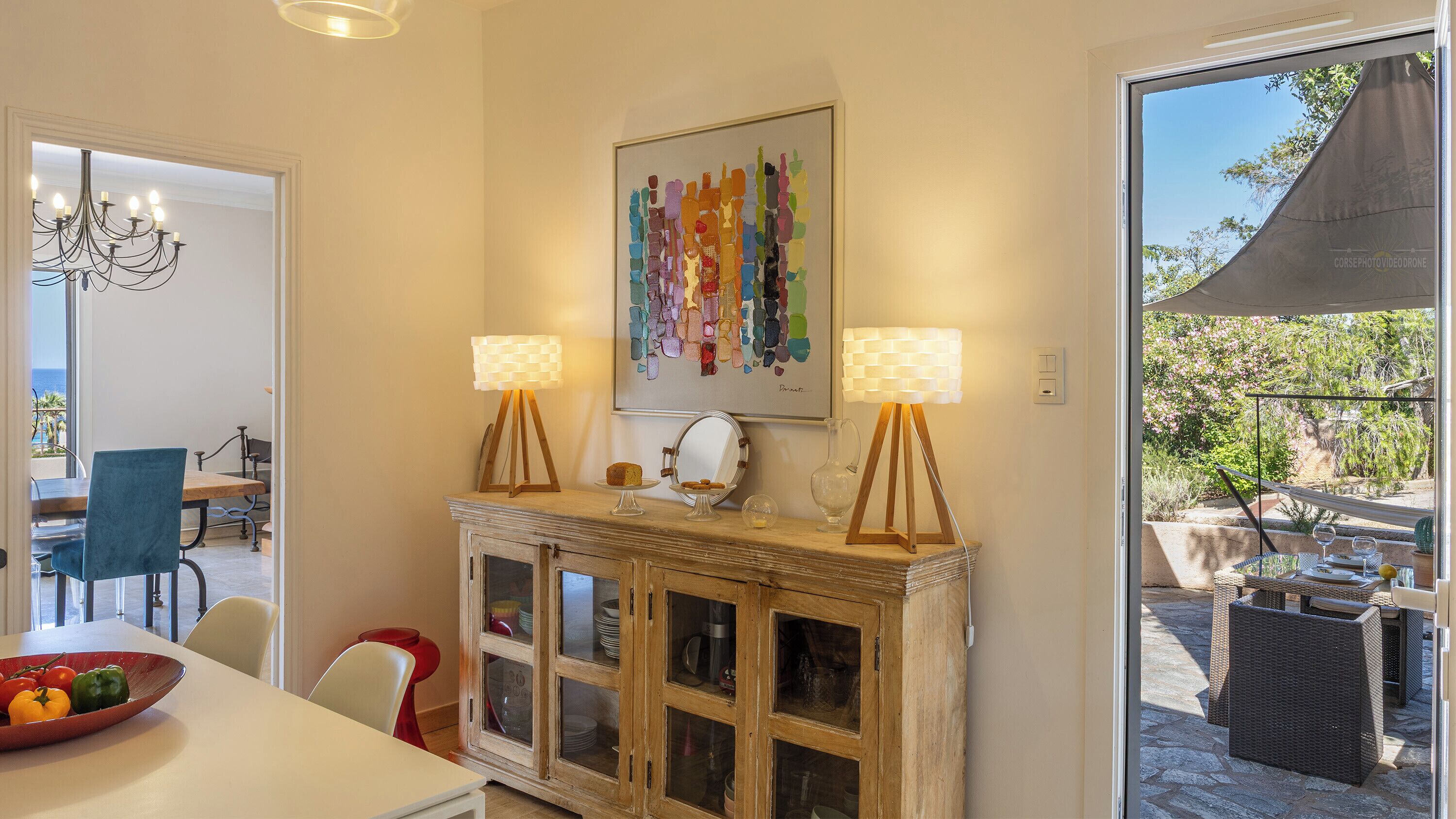 Uma área de refeições com mesa, cadeiras, um armário com portas de vidro, duas luminárias de chão e um quadro na parede.