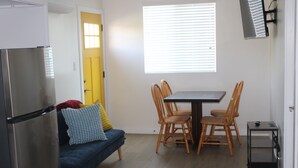 Una cocina compacta con refrigerador, una mesa de comedor con sillas, un sofá con cojines y una ventana con persianas.