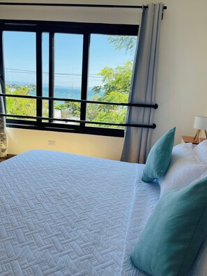 A bed with a quilt, a window with curtains, and a view of greenery and a body of water. - Perfect Place in Samana 1 (Samaná)