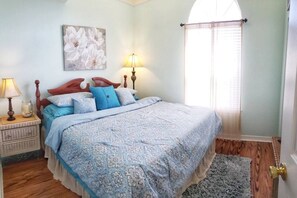 Ein Schlafzimmer mit einem Bett, Nachttischen, einer Lampe, einem Fenster mit Vorhängen und einem Blumenbild an der Wand.