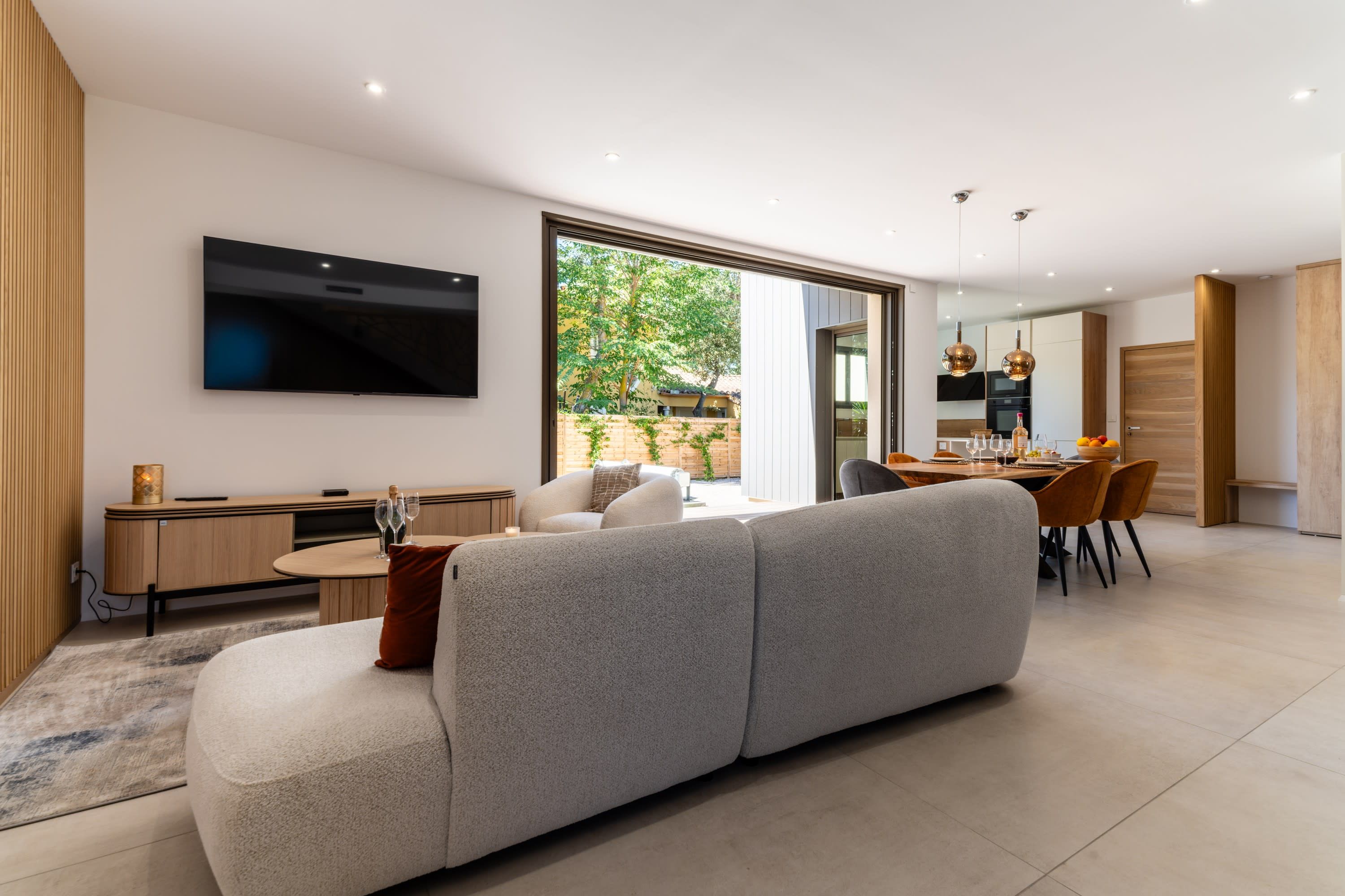 A modern living room with a large grey sofa, a flat-screen TV mounted on the wall, and a dining area with a wooden table and chairs.