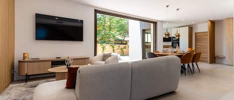 A modern living room with a large grey sofa, a flat-screen TV mounted on the wall, and a dining area with a wooden table and chairs.
