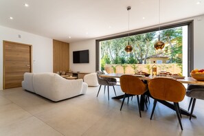 A modern living room with a sofa, dining table, chairs, and a large window with a view of greenery. - Villa Thétis • Beach, pool & good times (Agde)