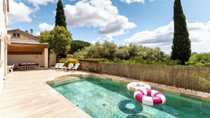 A clear swimming pool with lounge chairs and a wooden deck, surrounded by greenery and tall trees.