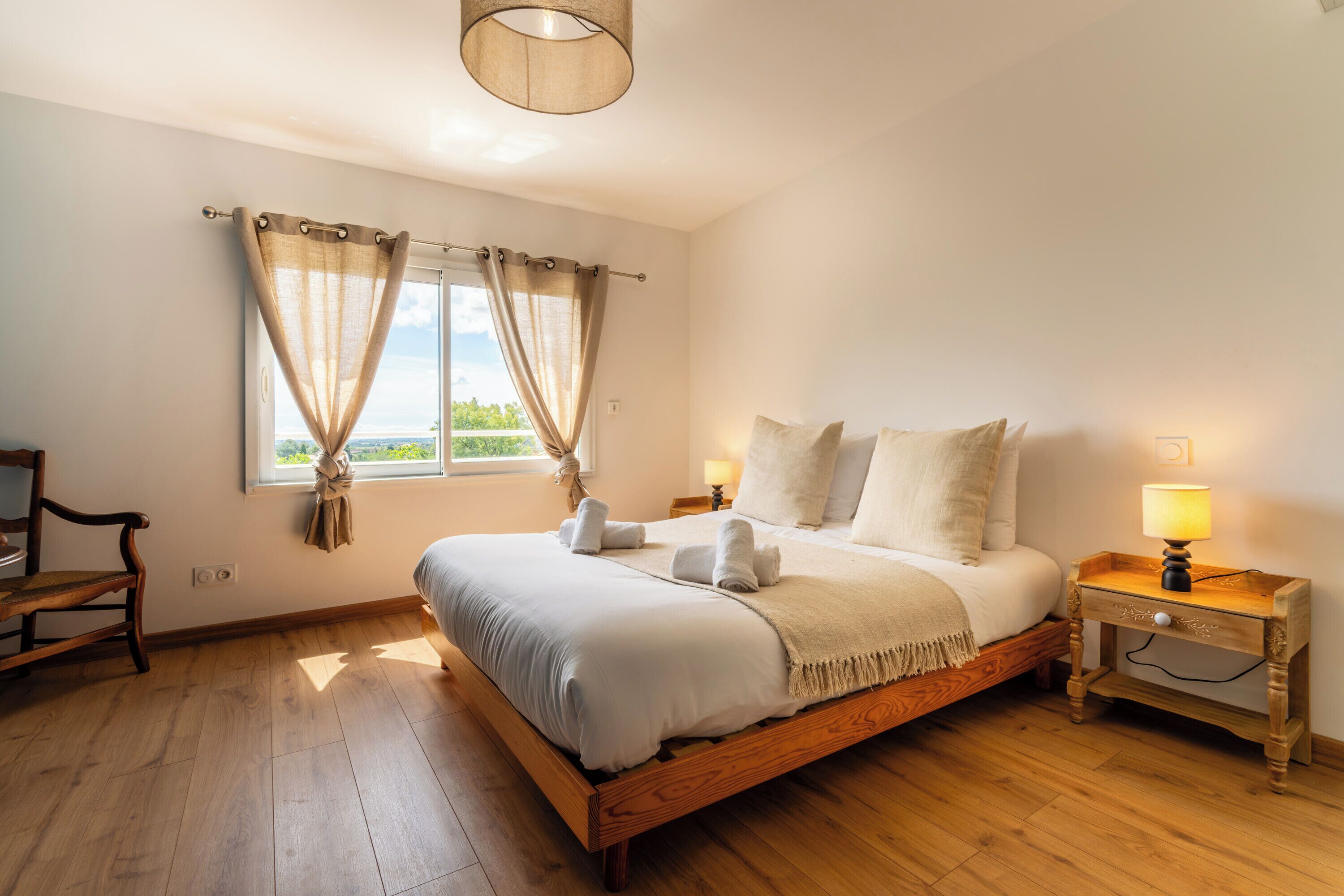 Een slaapkamer met een houten bed, nachtkastje met een lamp, een stoel en een raam met gordijnen.