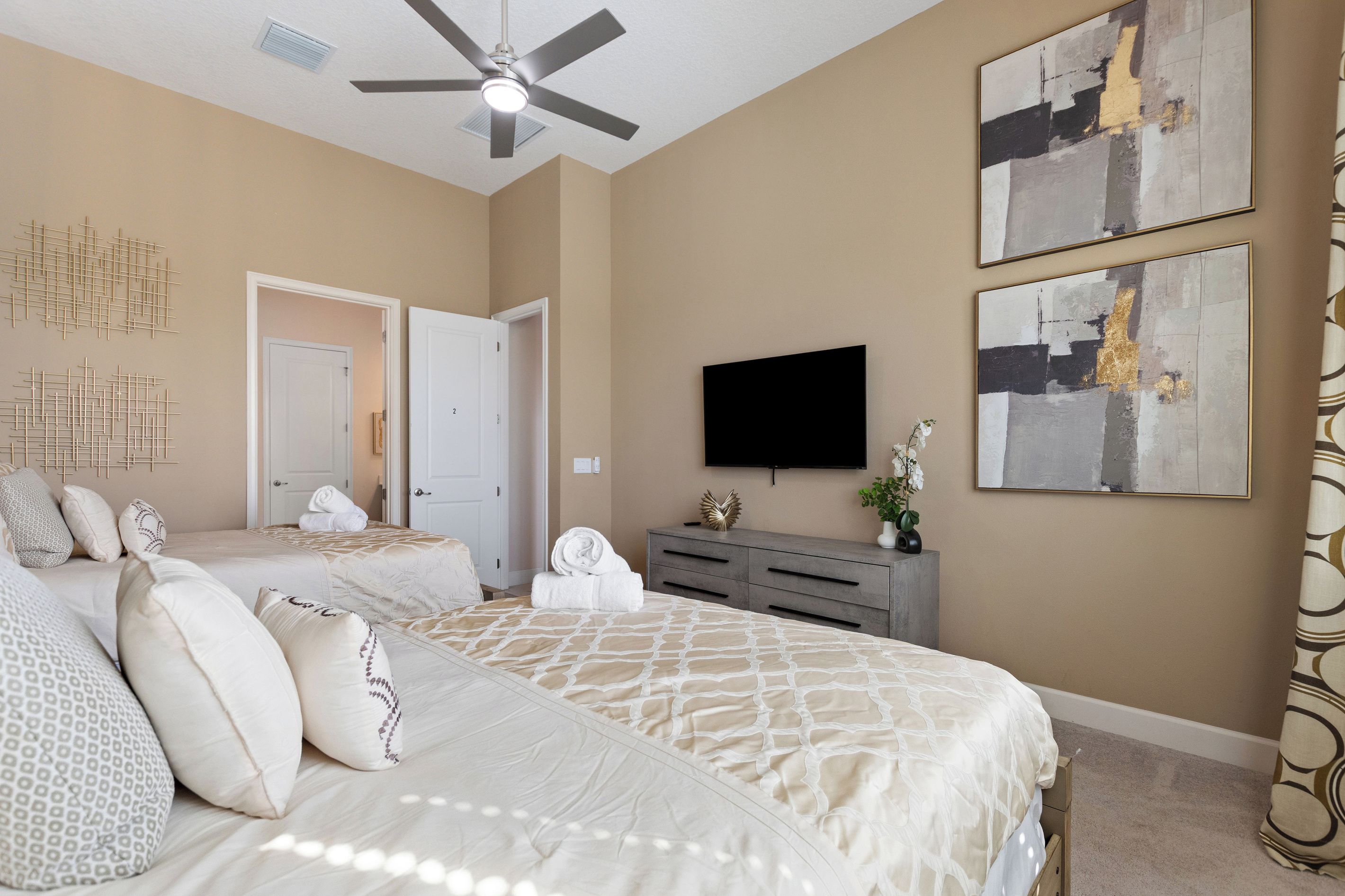 A hotel room with two beds, a ceiling fan, artwork on the walls, and a flat-screen TV.