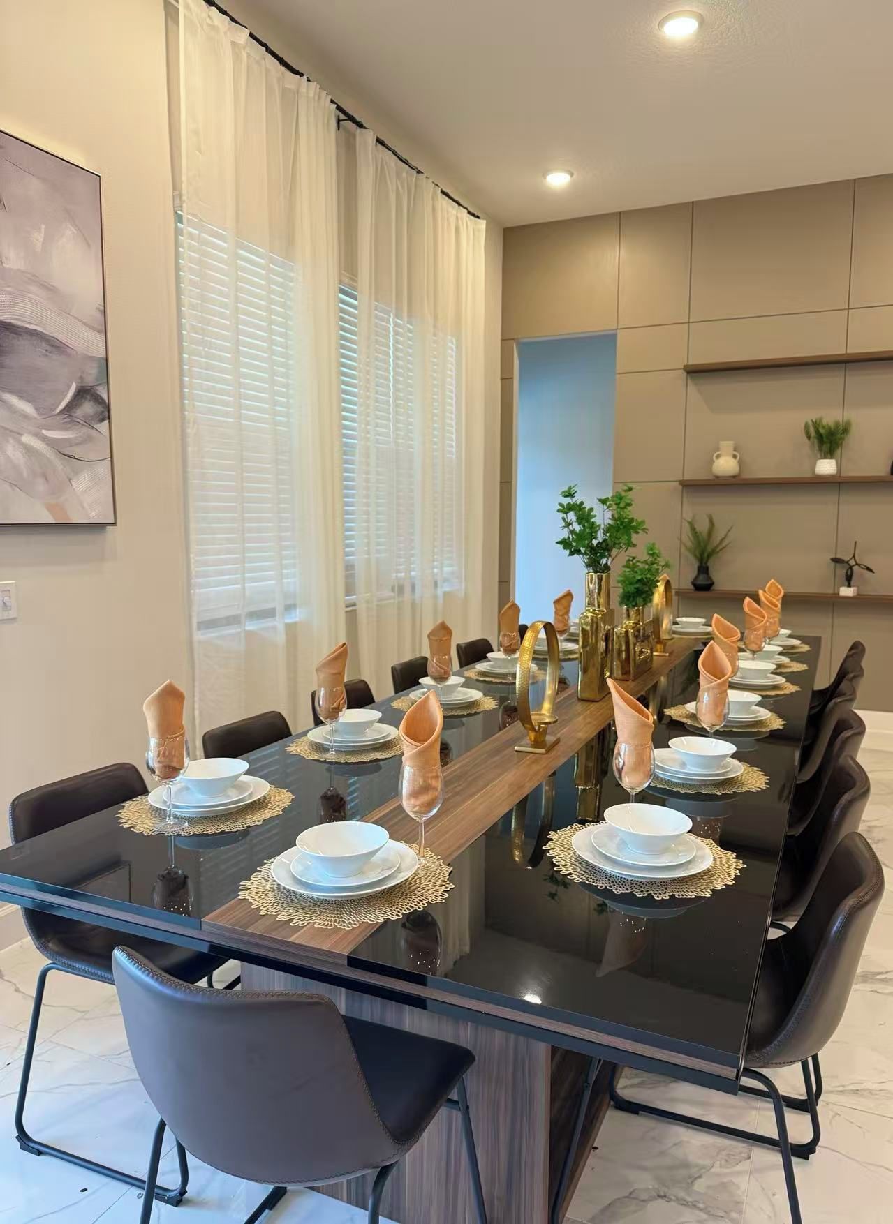 Une salle à manger moderne avec une longue table dressée pour un repas, dotée d’un plateau en verre, de chaises noires et d’éléments décoratifs dorés.