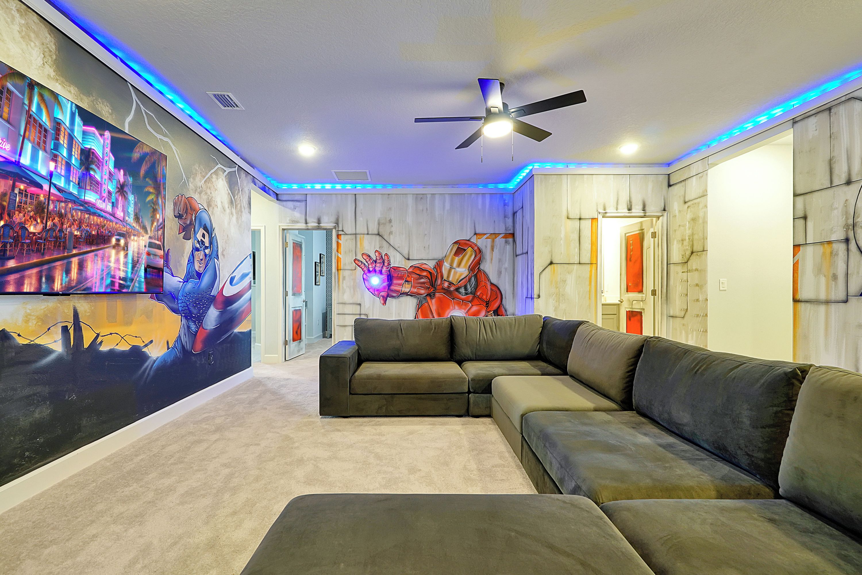 A room with a sectional sofa, a ceiling fan, and a wall mural featuring a superhero.