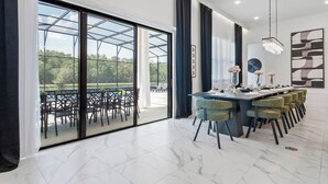 A modern dining area with a glass-enclosed patio, a long table with chairs, and a chandelier. - Regal 15BR 16.5BA, Sleeps 40, Lake View, Theater (Kissimmee)