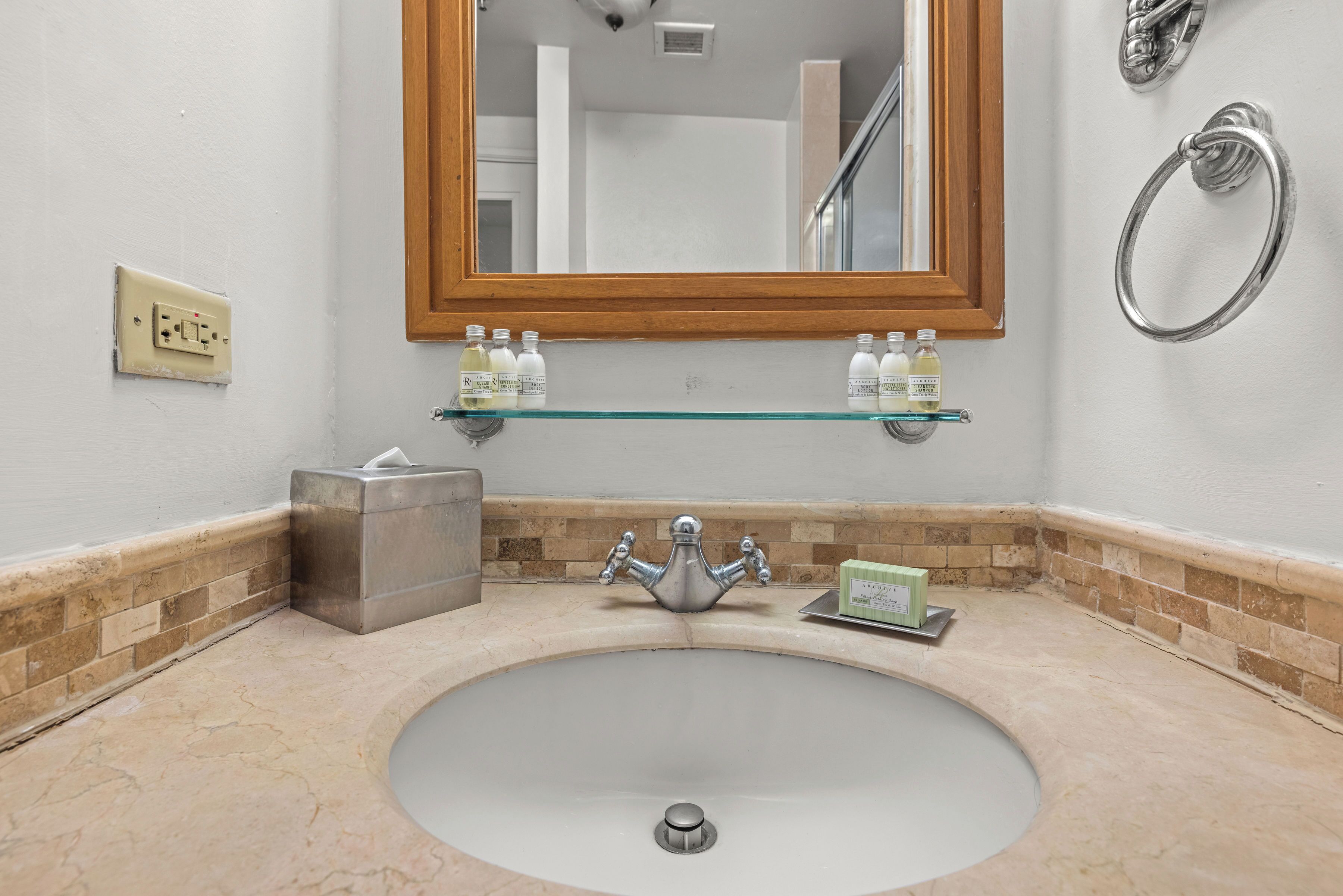 A bathroom with a sink, mirror, towel ring, and toiletries on a shelf.