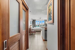 A hotel corridor with wooden doors, a bed with a nightstand, a chair, a ceiling fan, and a view of the city through large windows. - Condado Lagoon - Waterfront Escape (San Juan)