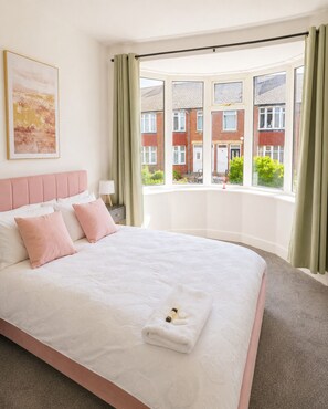 Ein Schlafzimmer mit einem großen Bett, einem Fenster mit Vorhängen, einem Nachttisch und einem Bild an der Wand.