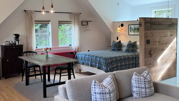 A loft-style room with a high ceiling, exposed beams, a dining area with a table and chairs, a sofa with patterned cushions, and a bed with a checkered duvet.