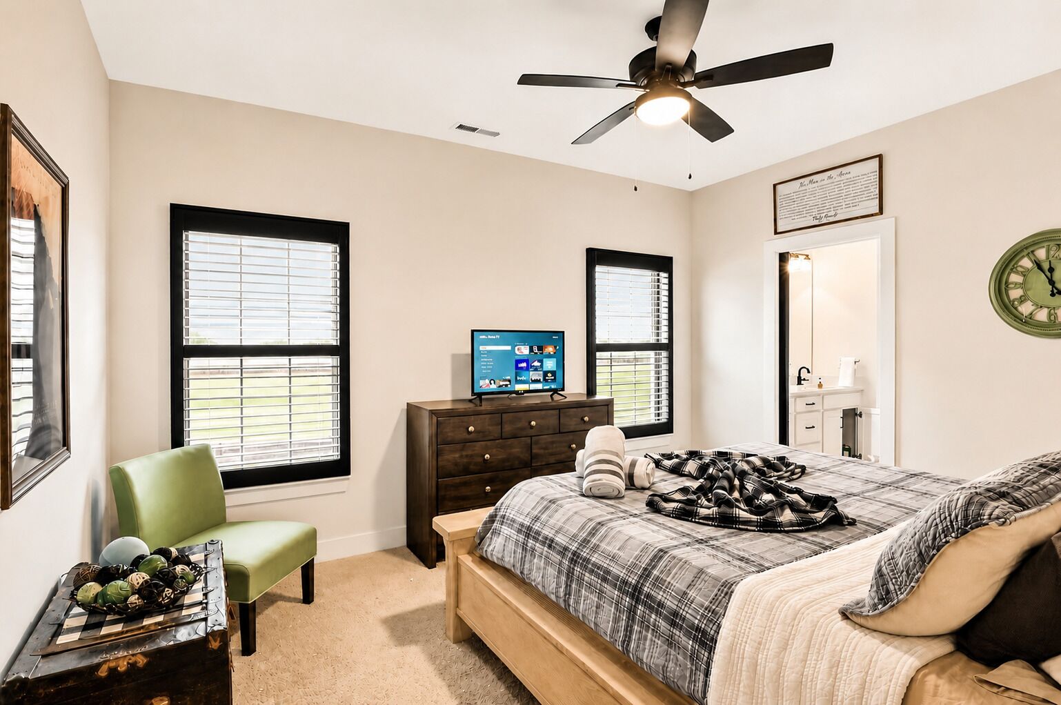 A bedroom with a bed, a desk, a chair, a dresser, a ceiling fan, and two windows.
