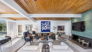 A modern living room with a wooden ceiling, a large abstract painting, a fireplace, and a flat-screen TV. - Luxury Resort Oasis | New Pool, Spa, Fire Pit, EV (Palm Springs)