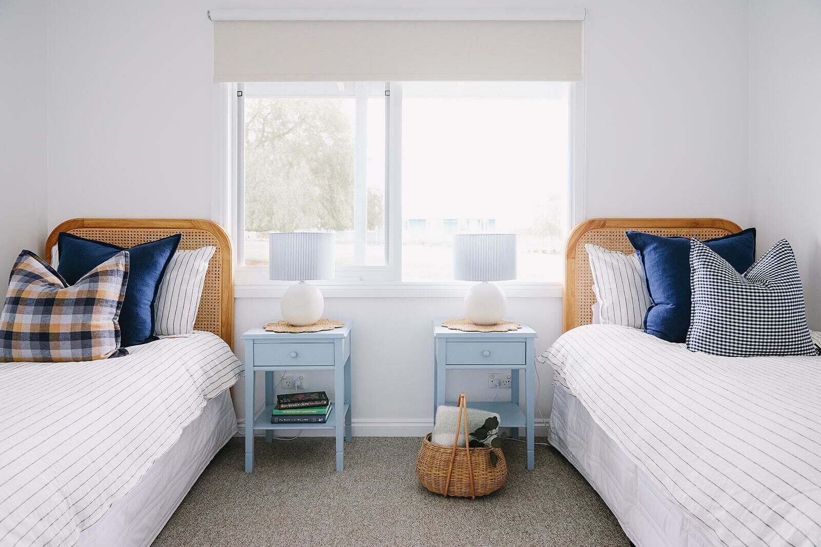 A bedroom with two beds, a window, bedside tables with lamps, and a basket on the floor.