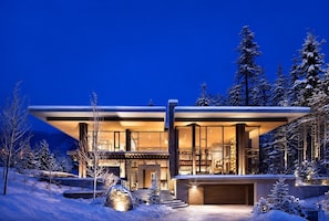Ein modernes, zweistöckiges Haus mit großen Fenstern, einer überdachten Terrasse und verschneiter Landschaft.