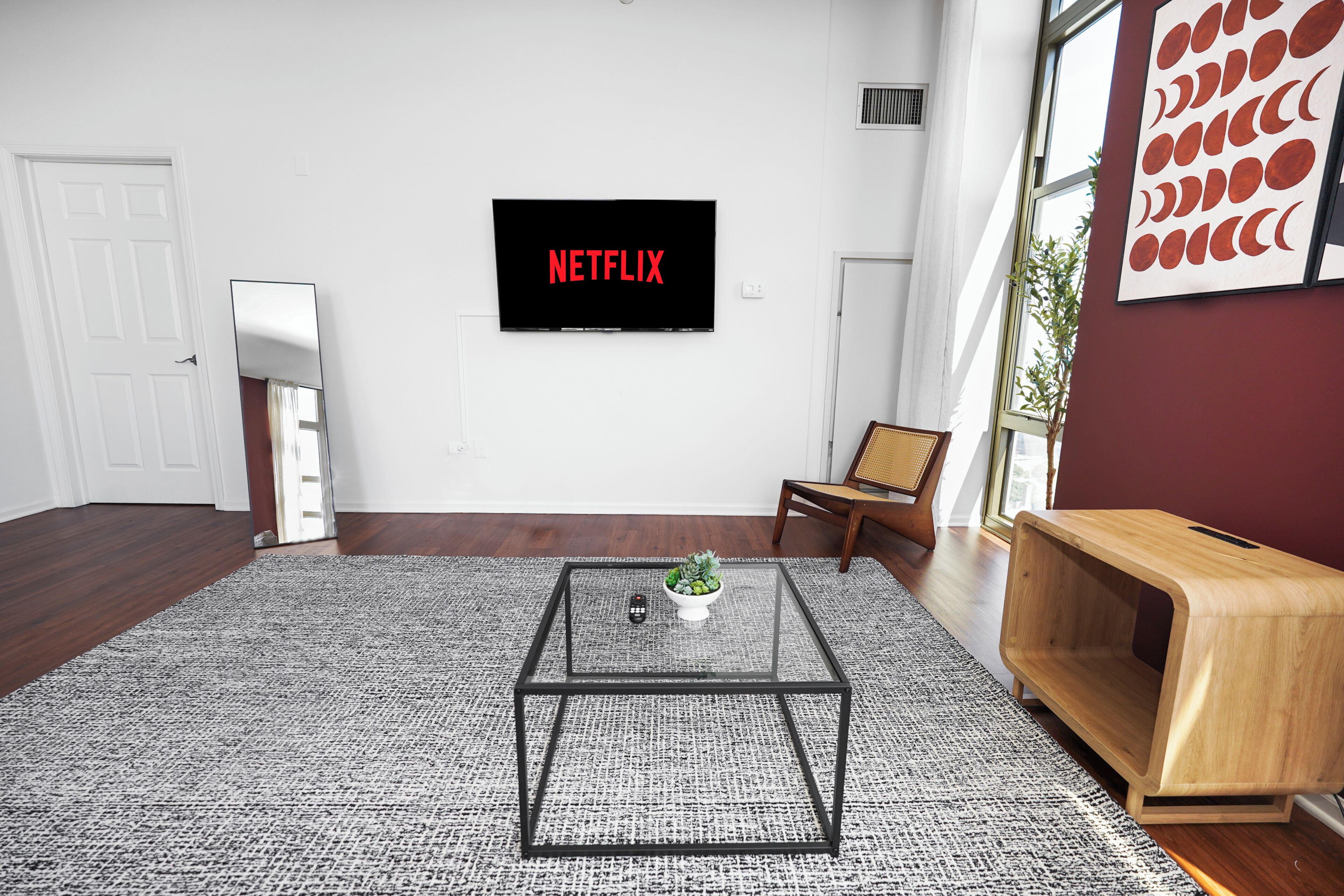 A modern living room with a flat-screen TV displaying 'NETFLIX,' a glass coffee table, a wooden side table, and a large window.