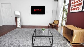 Ein modernes Wohnzimmer mit einem Flachbildfernseher, der 'NETFLIX' anzeigt, einem Glastisch, einem Holznachttisch und einem großen Fenster.