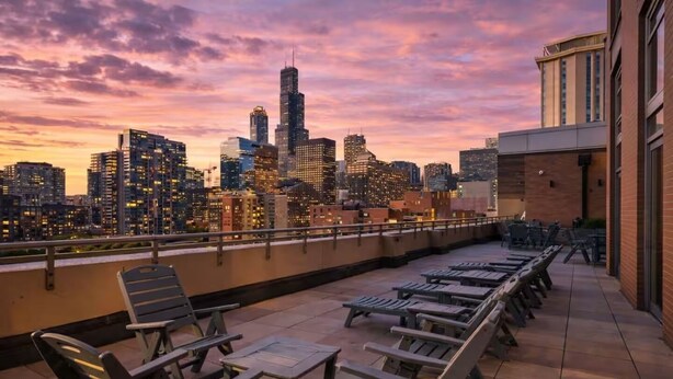 Eine Dachterrasse mit Lounge-Möbeln und Tischen mit Blick auf eine städtische Skyline bei Sonnenuntergang.