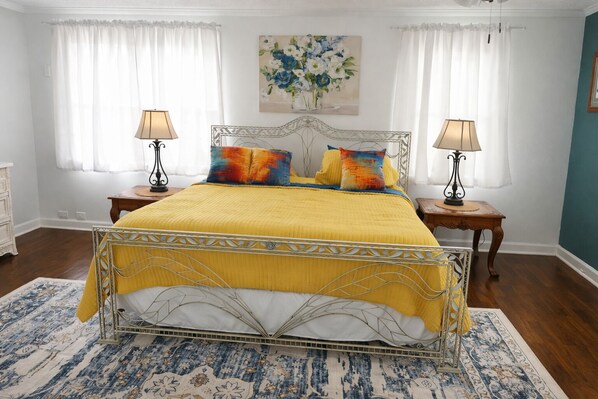 Un lit avec un cadre en métal, une couvre-lit jaune et des coussins colorés. Il y a deux tables de chevet avec des lampes. Un tableau représentant des fleurs est accroché au mur au-dessus du lit.