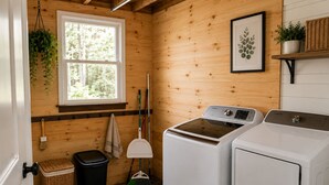 A laundry room with two washing machines, a broom, a trash bin, and a window.