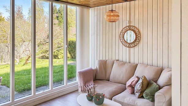 Ein Zimmer mit großem Fenster, einem beigen Sofa, einem kleinen runden Tisch und einer Pendelleuchte.