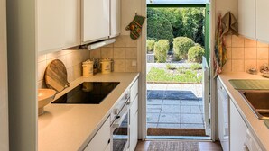 Eine Küche mit weißen Schränken, einem Holzstuhl und Blick auf einen Garten durch die Tür.
