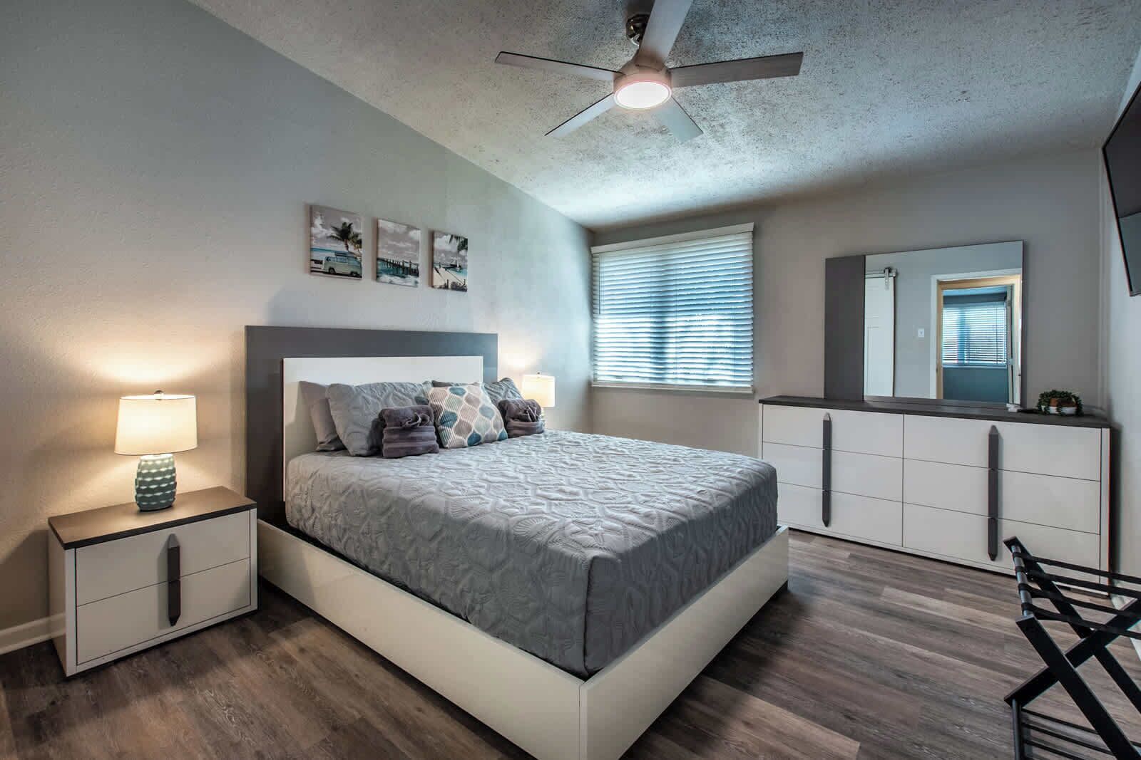 A bedroom with a bed, nightstands, a ceiling fan, a window with blinds, a dresser, and a mirror.