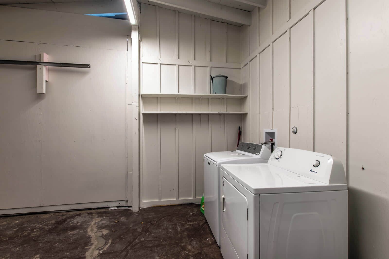 A laundry room with two white front-loading washers and dryers, a metal drying rack, and a small shelf with a bucket.