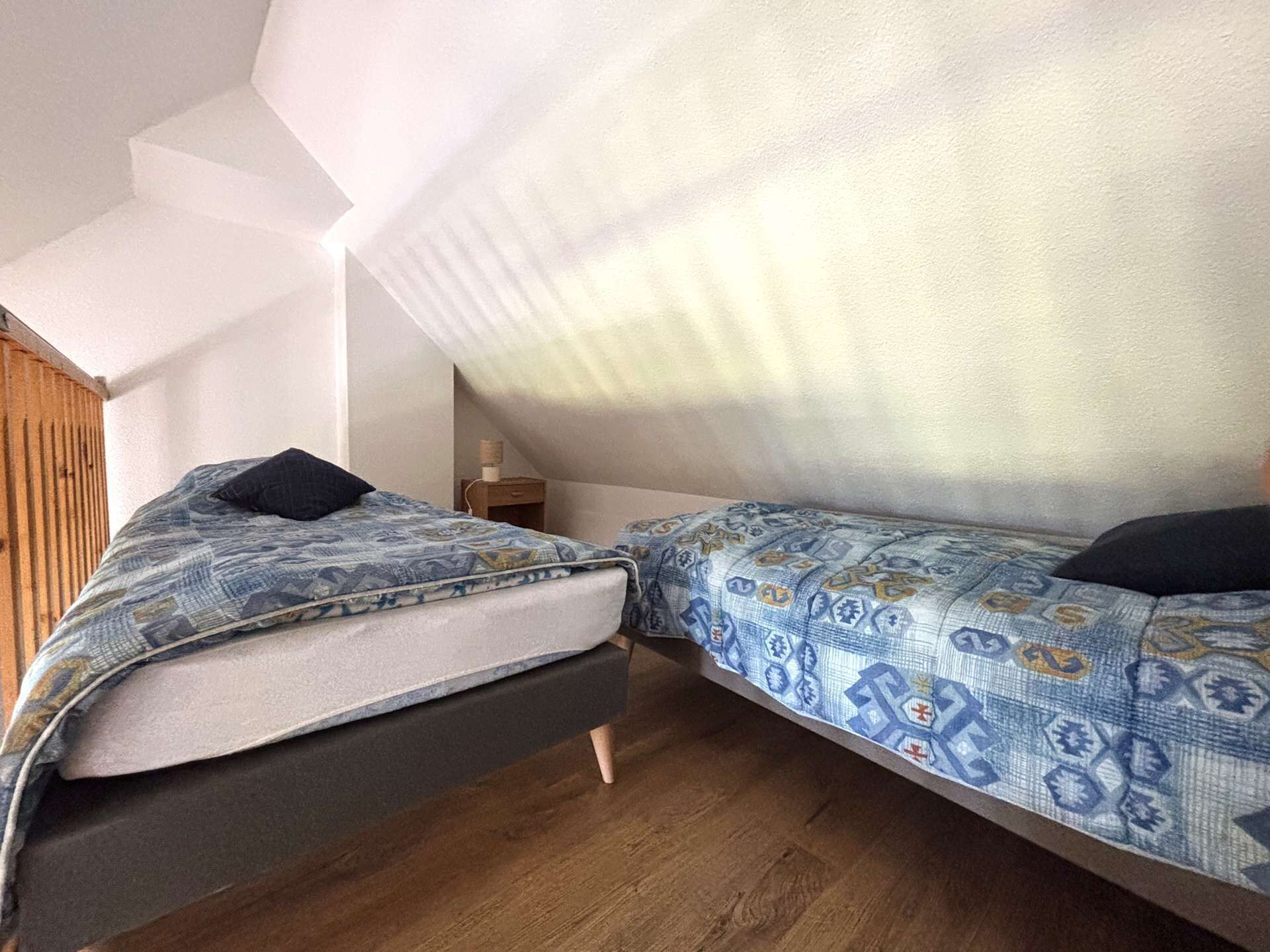 A small, cozy room with two beds, wooden flooring, and a sloped ceiling.