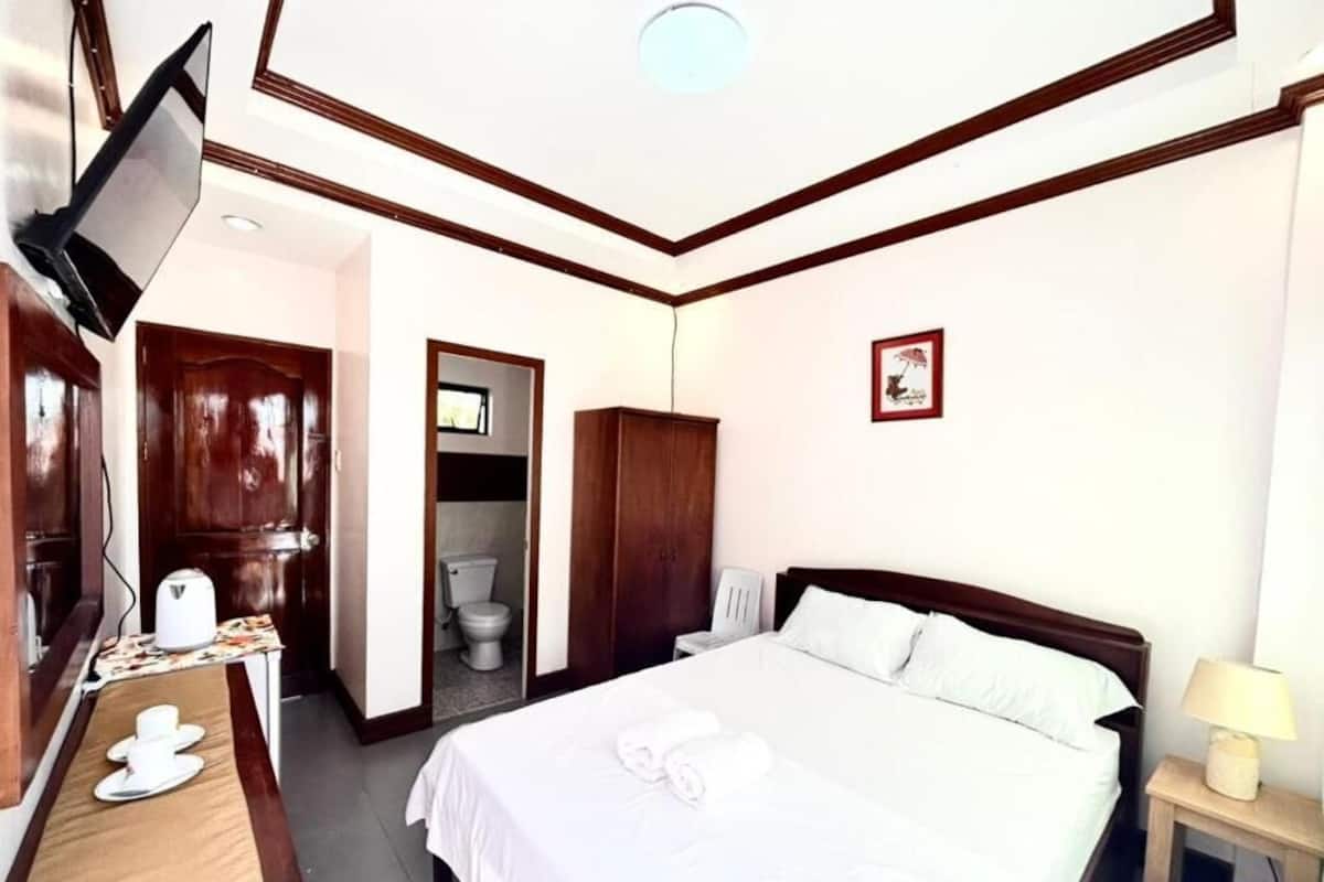 A hotel room with a bed, a desk with a chair, a television, and a bathroom visible through an open door.