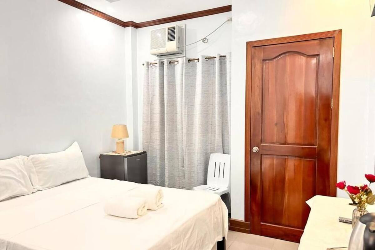 A bedroom with a bed, a nightstand with a lamp, a wooden door, and a curtain.