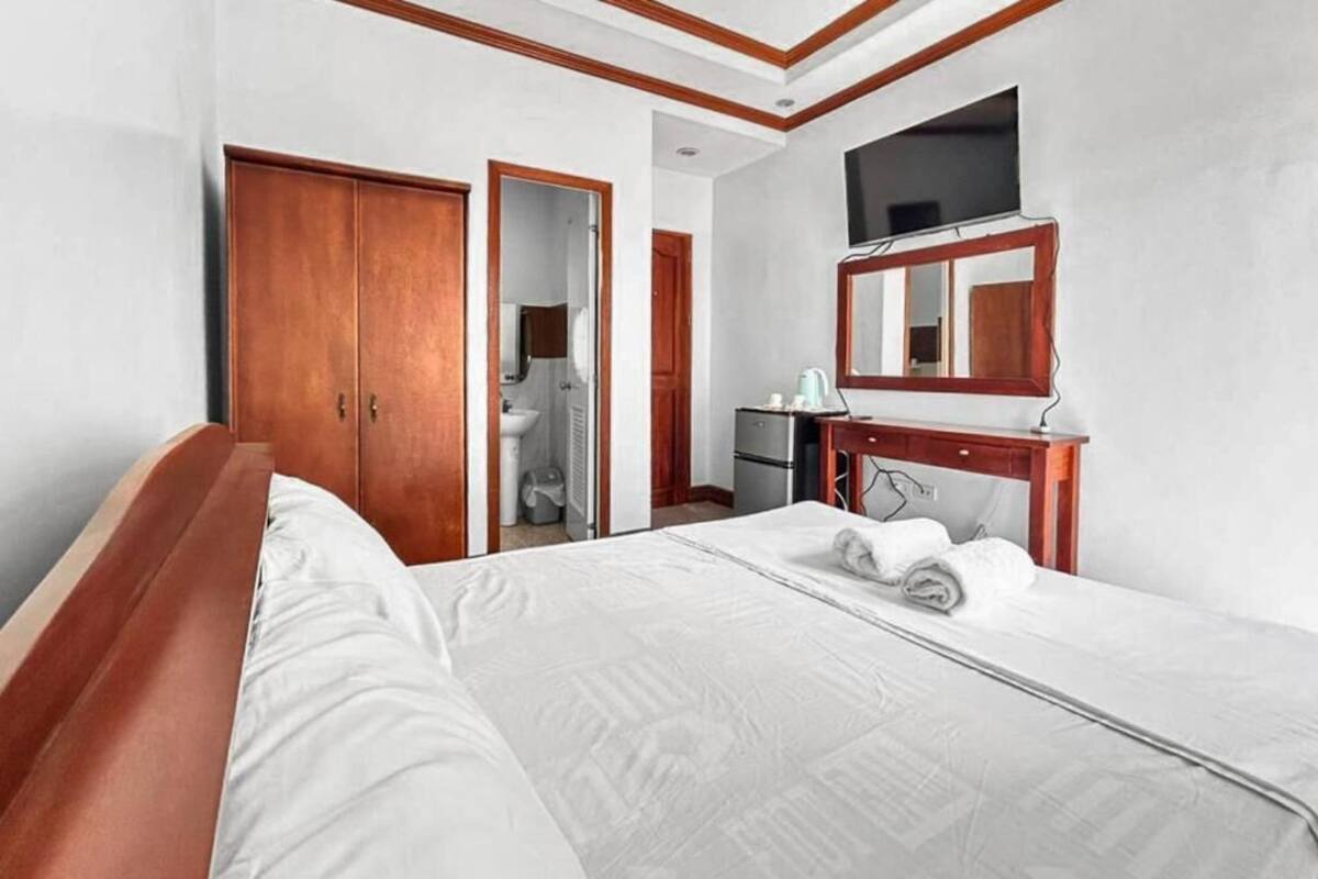 A hotel room with a bed, a wooden wardrobe, a desk with a mirror, a television, and a bathroom visible through an open door.