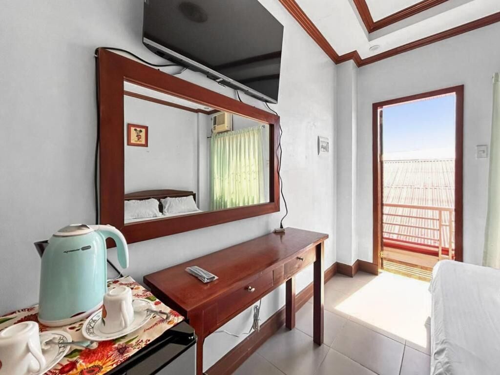 A hotel room with a wooden desk, a television mounted above it, a window with a view of a balcony, and a small table with a kettle and cups.
