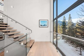 A modern interior with a glass staircase, a framed picture on the wall, and a view of snow-covered trees through large windows. - The Mountaintop | Kadenwood Ski In Ski Out | Views (Whistler)