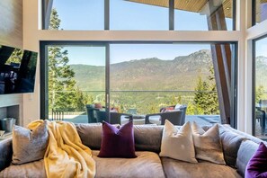 A modern living room with a large window overlooking a mountainous landscape, a sofa with cushions, and a flat-screen TV on the wall. - The Mountaintop at Kadenwood | Ski in Ski out (Whistler)