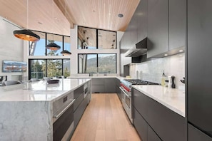 A modern kitchen with a large island, built-in appliances, and large windows offering a view of the outdoors. - The Mountaintop at Kadenwood | Ski in Ski out (Whistler)