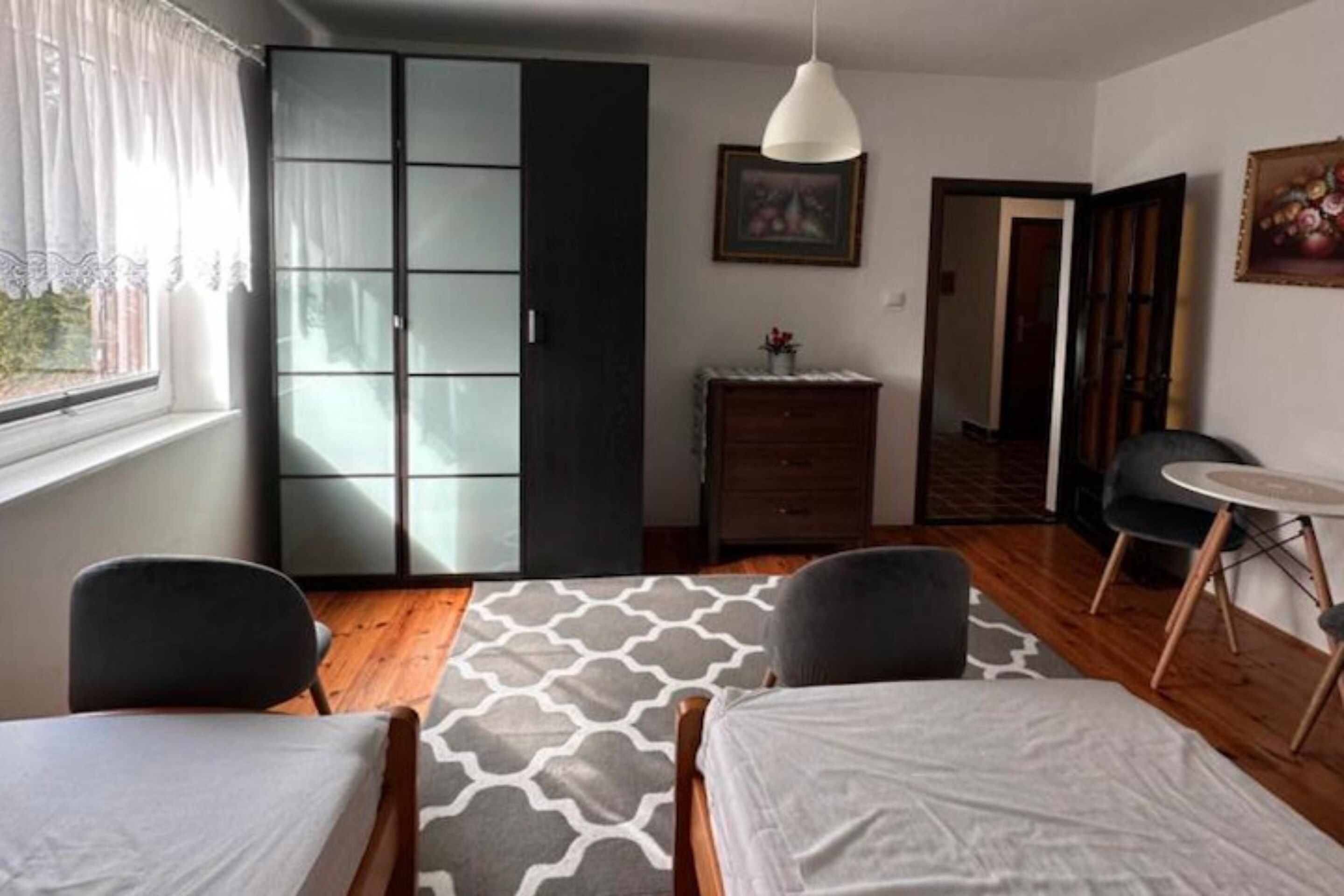 Quarto com cama, duas cadeiras, cômoda, janela com cortinas e um quadro na parede.