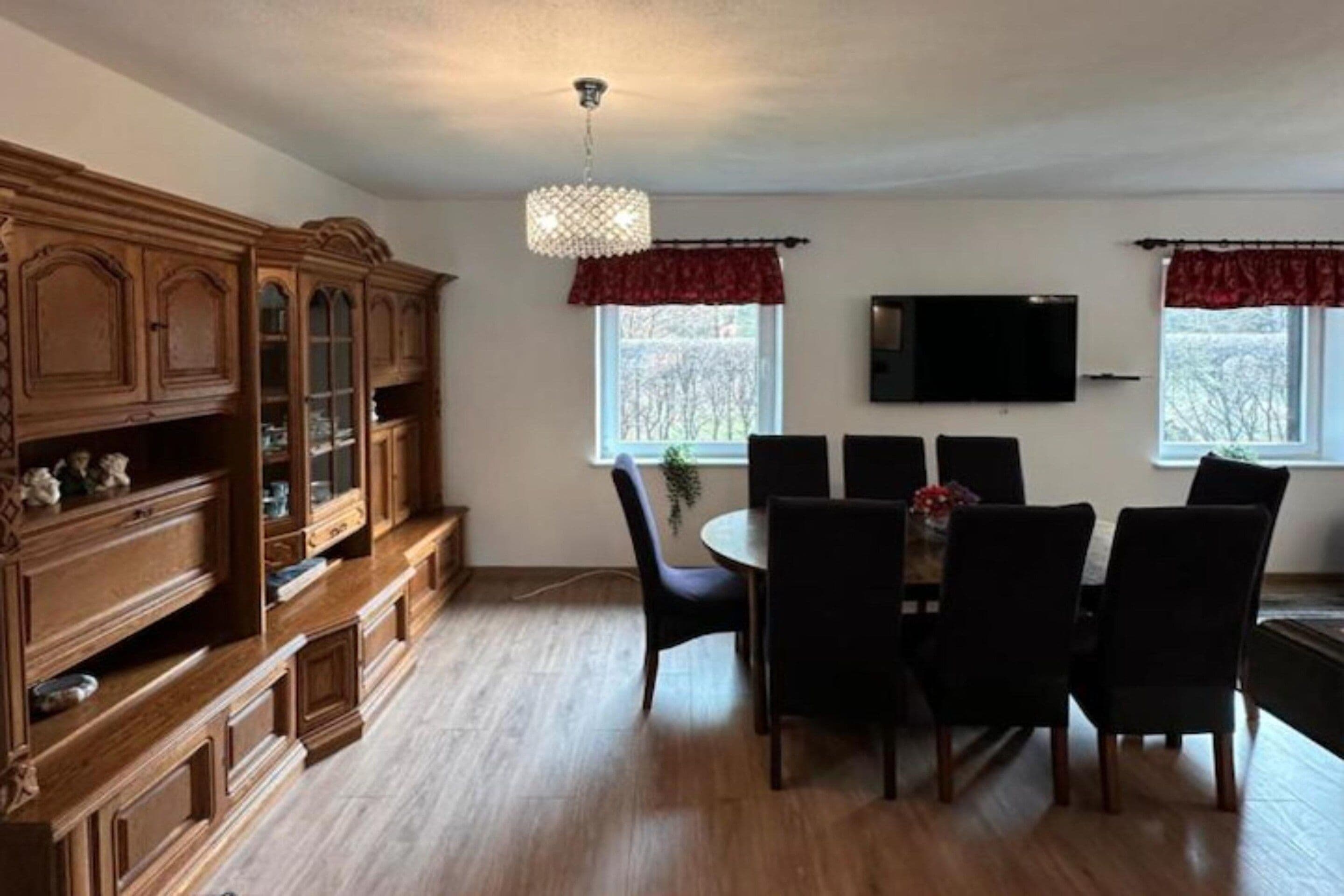 Uma área de jantar com um armário de madeira, um lustre, uma TV na parede e uma janela com vista para árvores.